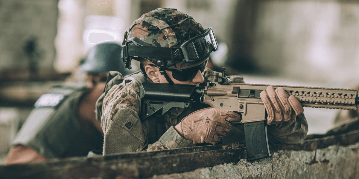Soldado equipado com uniforme camuflado, capacete e óculos táticos aponta um rifle durante operação militar em ambiente urbano.